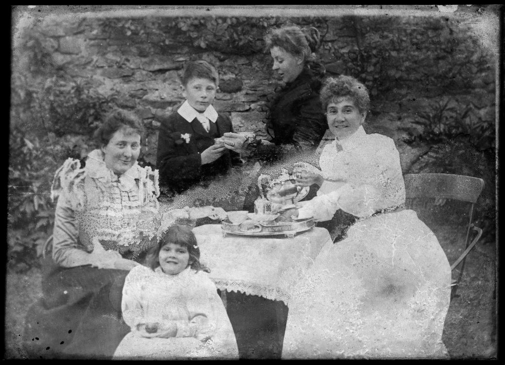 Three women with a boy and girl, all in late Victorian dress, seated around a tea table in a garden.