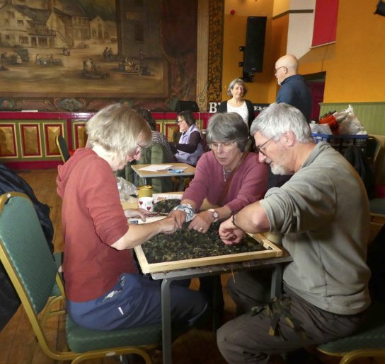 Three people work intently on a net on a small table in front of them. Behind them, another table can be seen, both in front of the fire screen at Settle Victoria Hall theatre