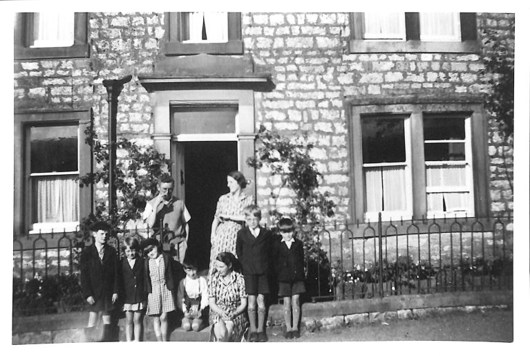 A black and white photo of seven children and two children outside a terraced house. They are posing as a group for the photo