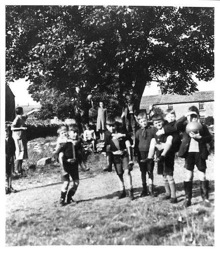 A black and white photo of around ten children running around under a tree in full leaf