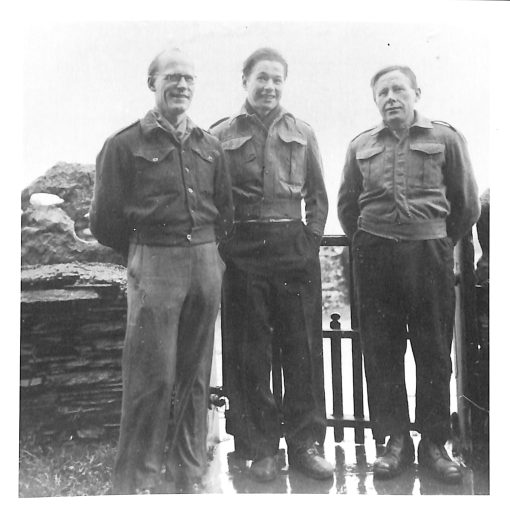 Three men smile for the camera in front of a farm gate. They are dressed in uniforms. The photo is black and white.