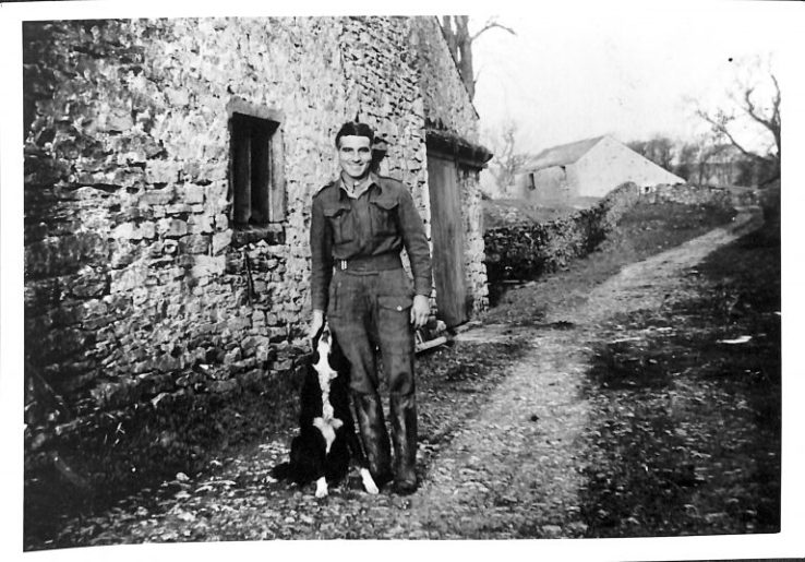 A black and white photo of a man in uniform outside a stone barn. By his side sits a border collie dog looking up at him.