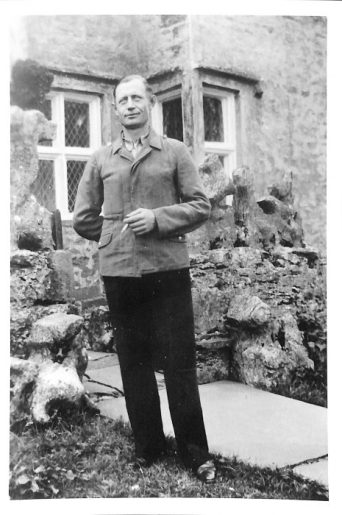 A black and white photo of a man in uniform jacket and black trousers, smoking a cigarette in front of a farmhouse