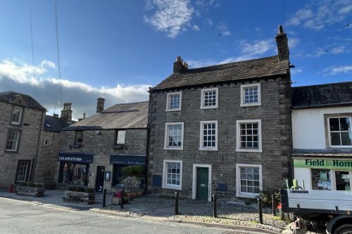 Colour photograph of a building. It has dark grey/brown stone bricks and eight windows - two ground floor, three middle, three upper - all surrounded by a pale frame. It is taller than the two buildings on either side, both of which are two stories high, one of which is white with a sign saying 'Field to Home' in green, and the other built from a similar grey/brown brick and a sign saying 'Car and Kitchen'. It is a sunny day with blue skies and some cloud.