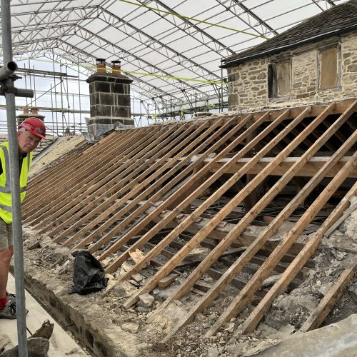 Man in a hard hat stands to the left of a roof stripped of its slates, showing the rafters