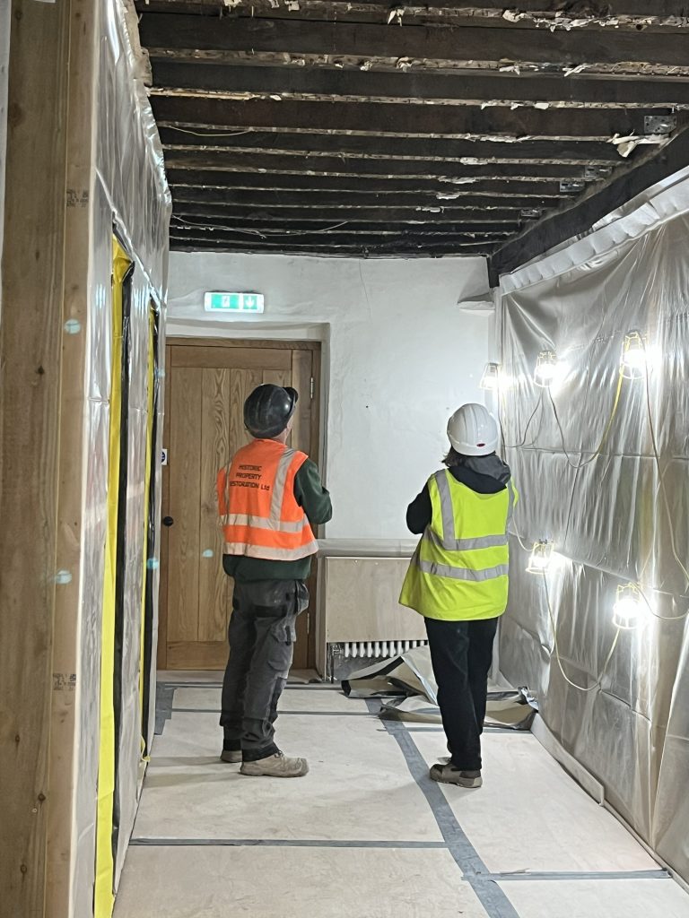 A man and a woman in hi-vis jackets inspect burnt joists in the ceiling above them