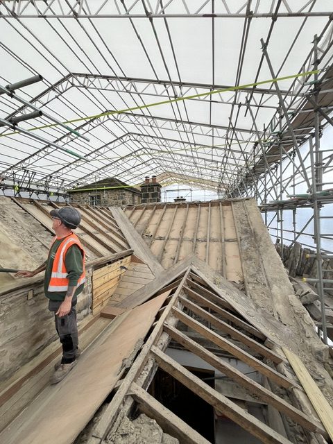 A builder in a hi-vis jacket stands on a partly repaired, complex seventeenth-century roof under a temporary roof covering
